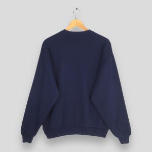 May include: A navy blue sweatshirt hangs from a wooden hanger. The sweatshirt has a crew neck and long sleeves. The fabric appears to be a soft, medium-weight material. The background is a solid, light gray color.