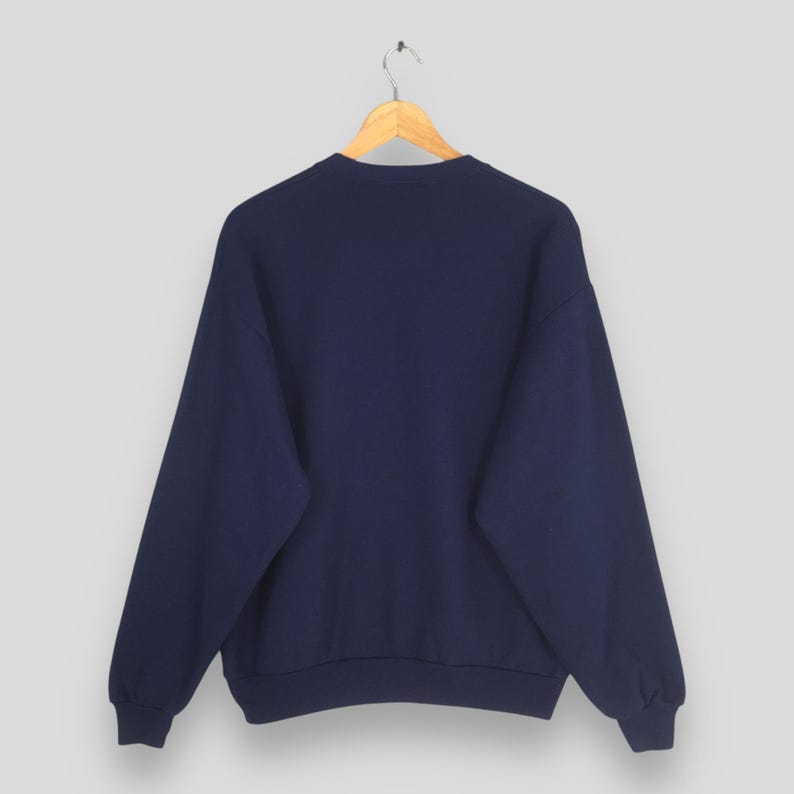 May include: A navy blue sweatshirt hangs from a wooden hanger. The sweatshirt has a crew neck and long sleeves. The fabric appears to be a soft, medium-weight material. The background is a solid, light gray color.