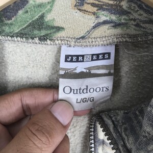 Puede incluir: Primer plano de una sudadera con estampado de camuflaje y cremallera. La etiqueta dice "JERZEES Outdoors L/G/G". La tela tiene una mezcla de tonos verdes, marrones y beige. La cremallera es visible en la parte inferior.