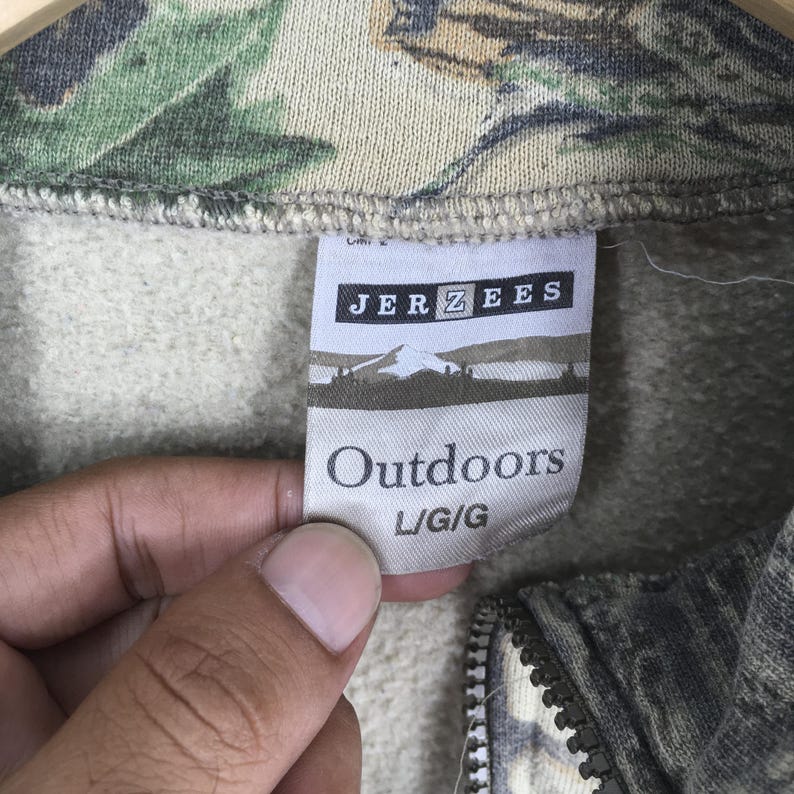 Puede incluir: Primer plano de una sudadera con estampado de camuflaje y cremallera. La etiqueta dice "JERZEES Outdoors L/G/G". La tela tiene una mezcla de tonos verdes, marrones y beige. La cremallera es visible en la parte inferior.