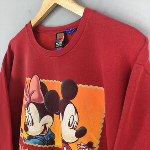 May include: Red crewneck sweatshirt featuring a graphic of Mickey and Minnie Mouse. The graphic is set against an orange background with a scalloped border. The sweatshirt has a "Mickey Unlimited" tag.