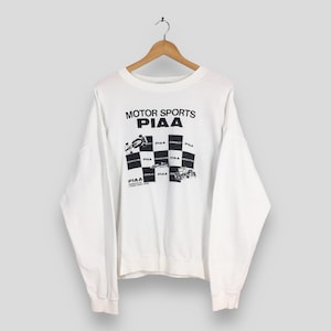 May include: White long-sleeve sweatshirt with "MOTOR SPORTS PIAA" printed in black. The design includes a checkered flag and race car graphics. The sweatshirt is hanging on a wooden hanger.