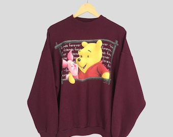 Op de afbeelding: Een bordeauxrood sweatshirt met een afbeelding van Winnie de Poeh en Biggetje. De afbeelding is omringd door de tekst "Friends forever" die meerdere keren wordt herhaald.