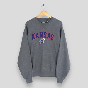 May include: A grey crew neck sweatshirt featuring the word "KANSAS" in red and blue arched lettering. Below the word "KANSAS" is a small cartoon Jayhawk mascot. The sweatshirt is suspended from a wooden hanger.