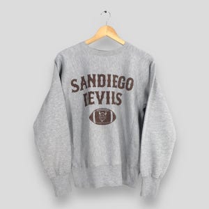 Op de afbeelding: Een grijze sweatshirt met de tekst "SANDIEGO DEVILS" in bruine letters boven een bruine voetbalafbeelding. De sweatshirt hangt aan een houten hanger.