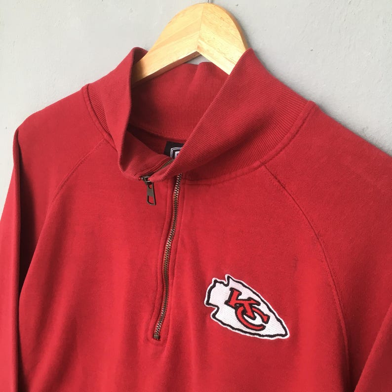 Op de afbeelding: Een rood sweatshirt met een kwartritssluiting, een hoge kraag en een metalen rits. Een geborduurde patch op de linkerborst toont een witte pijlpuntvorm met een zwarte omlijning en de rode letters KC in het midden van het ontwerp.