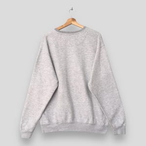Op de afbeelding: Een lichtgrijs sweatshirt met een ronde hals en lange mouwen, hangend aan een houten hanger. De sweatshirt heeft een geribbelde kraag, manchetten en zoom. De achterkant toont een eenvoudig, casual ontwerp.