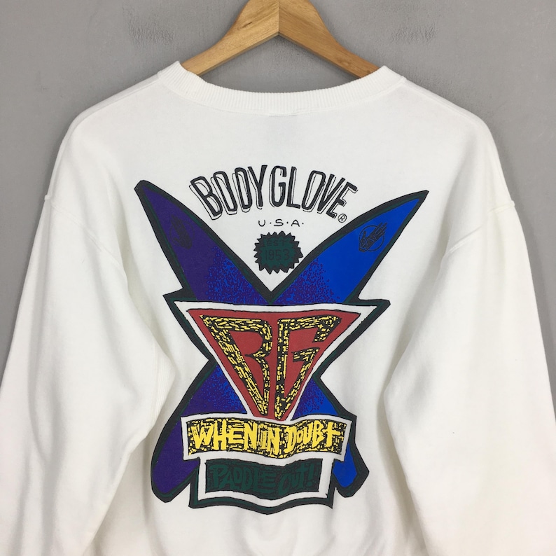 Op de afbeelding: Witte sweatshirt met een grafisch ontwerp met de tekst "BODYGLOVE U.S.A." en een kleurrijk logo. Het logo bevat blauwe en paarse vormen, een rode en gouden driehoek en de tekst "WHEN IN DOUBT PADDLE OUT."