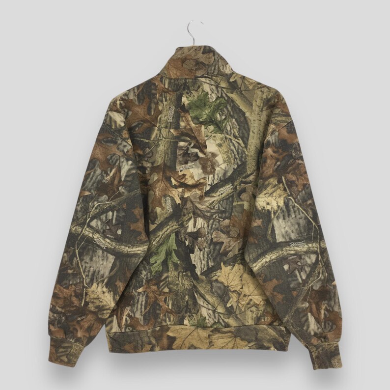 May include: A camouflage-patterned jacket with a high collar and a zipper closure. The jacket features a blend of brown, green, and tan hues, mimicking a forest environment. The sleeves have ribbed cuffs, and the jacket is hanging on a metal hanger.