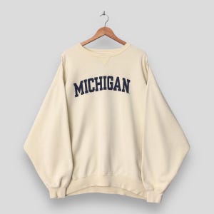 May include: A cream-colored crewneck sweatshirt featuring the word MICHIGAN printed in dark navy blue block letters across the chest. The garment has long sleeves, a ribbed collar, and a small triangular stitched detail below the neckline.