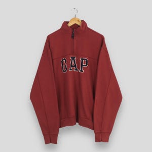 May include: A maroon Gap sweatshirt with a quarter-zip neckline. The word "GAP" is embroidered across the chest in navy blue letters with a white outline. The sweatshirt is hanging on a wooden hanger.