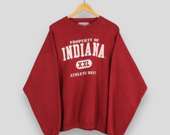 Może przedstawiać: Czerwona bluza XXL z napisem "PROPERTY OF INDIANA" w białych literach. Poniżej napis "XXL" w owalu i "ATHLETIC DEPT" w kolorze białym. Bluza wisi na drewnianym wieszaku.
