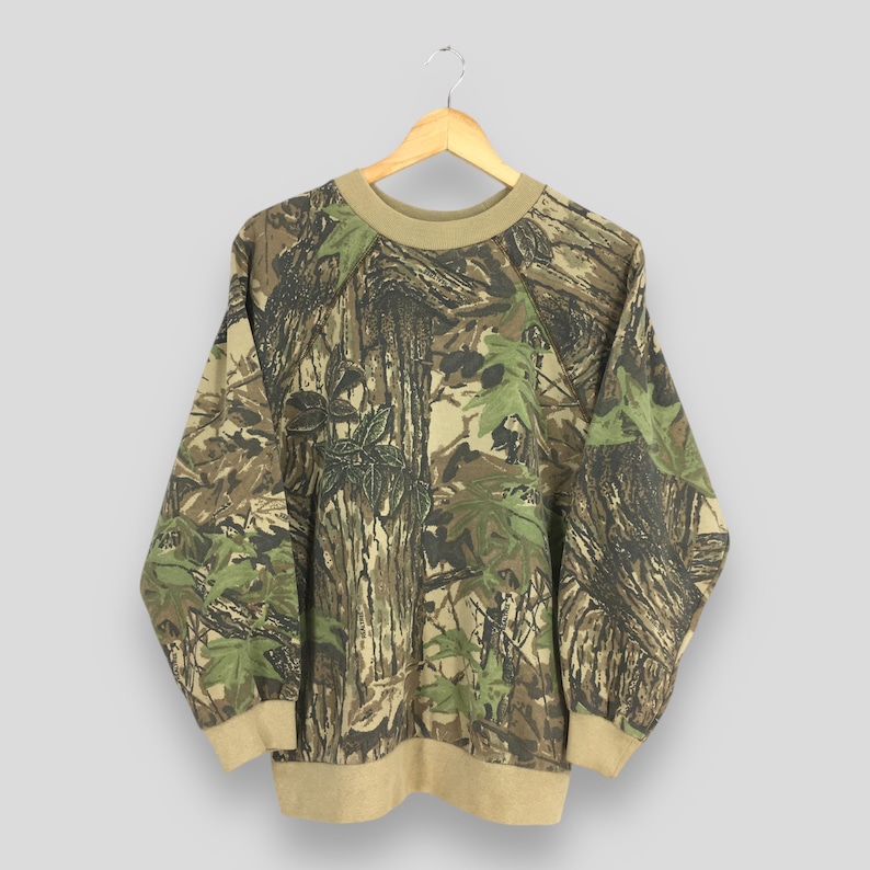 Op de afbeelding: Een sweatshirt met camouflagepatroon met een ronde hals en lange mouwen. Het ontwerp heeft een mix van bruine, groene en bruine kleuren, die lijken op een boslandschap. De kraag, manchetten en zoom zijn effen bruin van kleur. Het sweatshirt hangt aan een houten hanger.