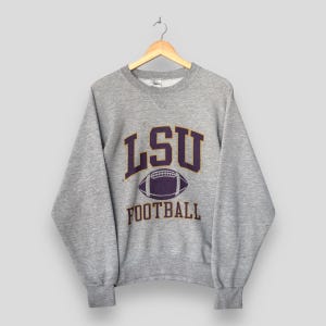 以下が含まれることがあります： 紫と金色の「LSU FOOTBALL」の文字が入ったヘザーグレーのスウェットシャツ。デザインは、紫色のフットボールのグラフィックの上に「LSU」の文字、下に「FOOTBALL」が配置されています。スウェットシャツはクルーネックで長袖です。