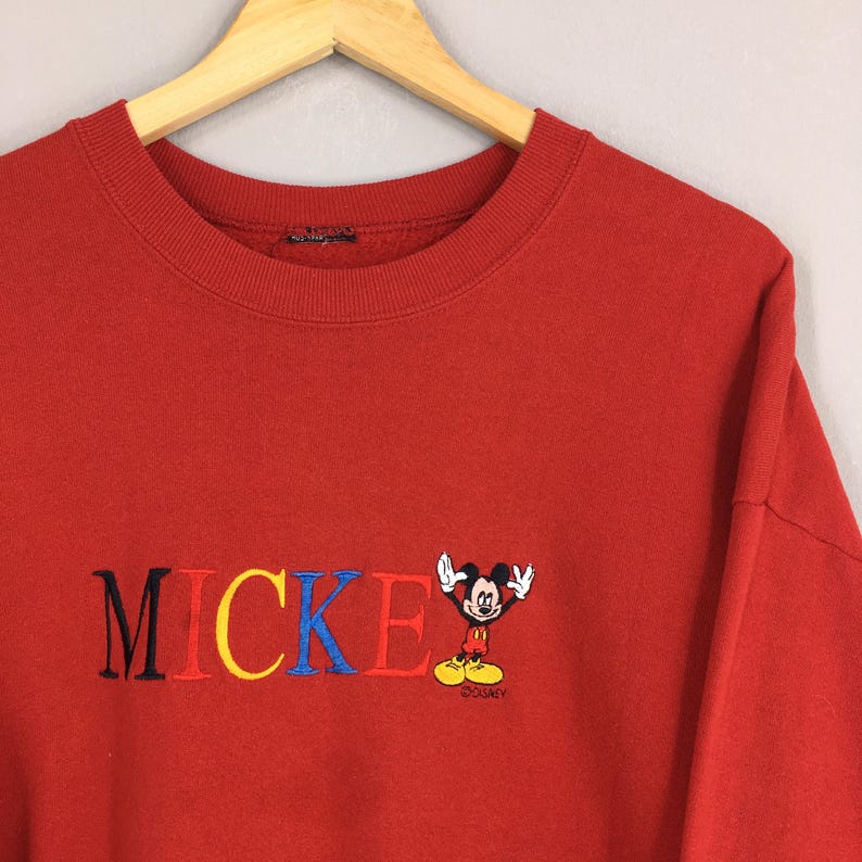 Peut inclure: Sweat-shirt rouge &agrave; col rond avec le mot "MICKEY" brod&eacute; en noir, jaune et bleu. Une petite image brod&eacute;e de Mickey Mouse est sur le c&ocirc;t&eacute; droit. Le sweat-shirt est suspendu &agrave; un cintre en bois.