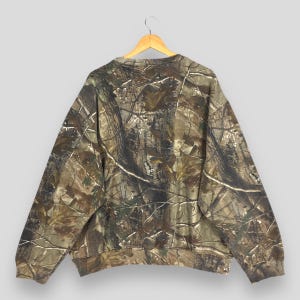May include: A camouflage-patterned sweatshirt hangs on a wooden hanger. The long-sleeved top features a brown, green, and tan woodland camouflage design. The sweatshirt is made of a soft material and has a crew neck.