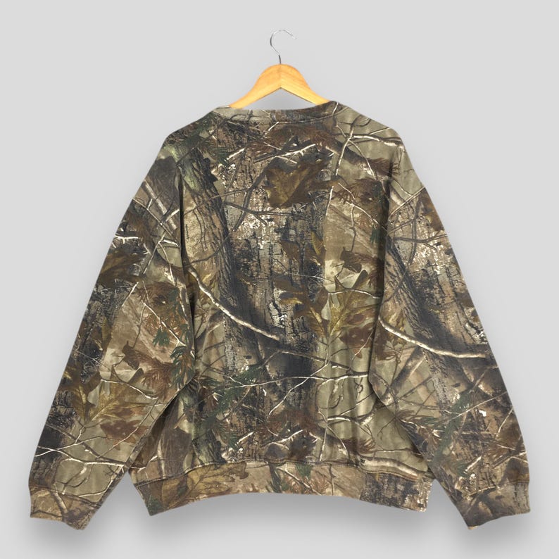 May include: A camouflage-patterned sweatshirt hangs on a wooden hanger. The long-sleeved top features a brown, green, and tan woodland camouflage design. The sweatshirt is made of a soft material and has a crew neck.