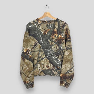 May include: A long-sleeved camouflage sweatshirt hangs from a wooden hanger. The pattern features shades of brown, green, and gray, mimicking a forest environment. The sweatshirt is made of a soft material and has a crew neck.