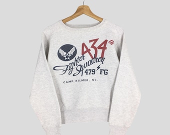 May include: A pale grey sweatshirt featuring the text "A34th Fighter Squadron 479 FG CAMP KILMER, NJ." in navy and red. The design includes a circular emblem with a star and wings.
