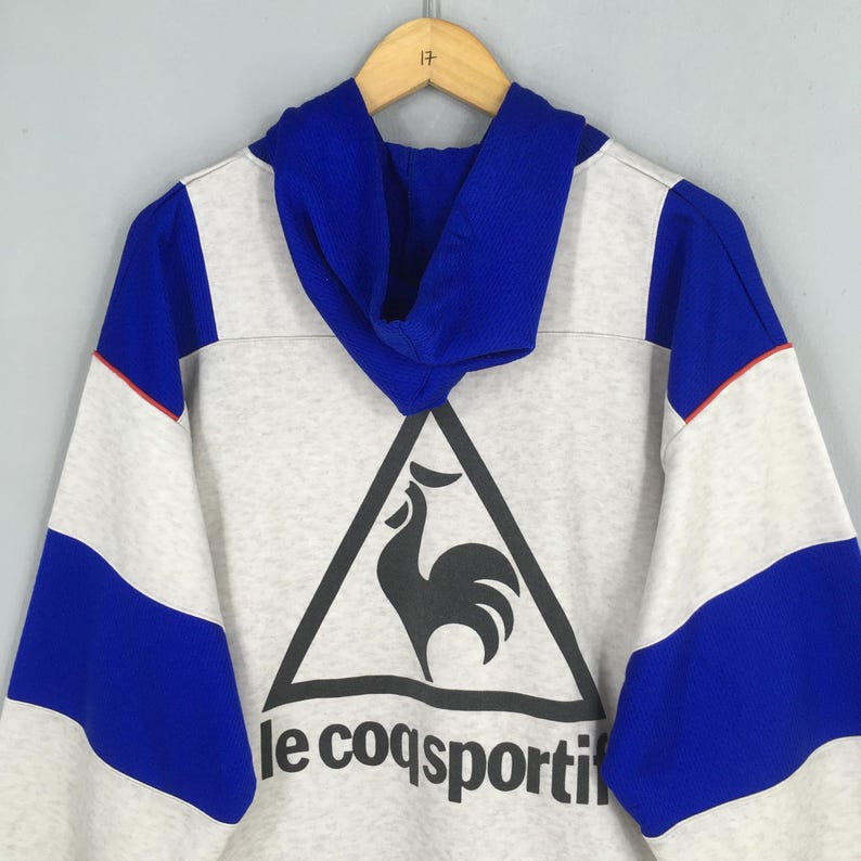 May include: A gray hoodie with blue and white accents. The hoodie features a large black triangle with a rooster logo and the text "le coq sportif" below it. The sleeves are blue and white with a red stripe.