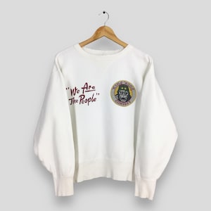 May include: White sweatshirt with the text "We Are The People" in maroon script. A circular patch features a gorilla with a military helmet and the words "King Kong Company". The sweatshirt is hanging on a wooden hanger.