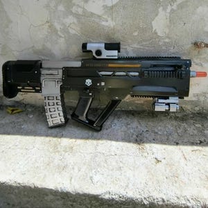 Puede incluir: Un rifle de juguete negro y plateado con diseño bullpup, que incluye una mira montada, un cargador extraíble y un emblema de calavera blanca en el lateral. La punta del cañón es de color naranja brillante y el cuerpo tiene empuñaduras texturizadas.