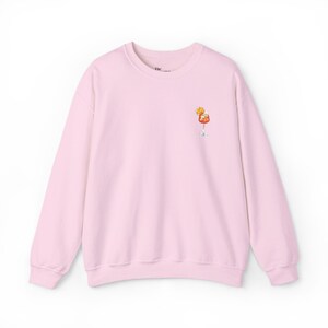 May include: Pale pink sweatshirt with a crew neck and long sleeves. A small cocktail graphic in a stemmed glass is on the front. The cocktail features orange and yellow detailing.