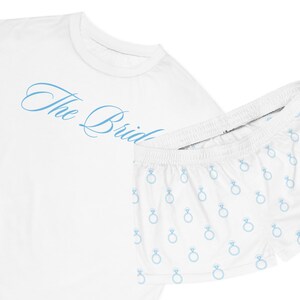 Puede incluir: Camiseta blanca con el texto "The Bride" en escritura azul claro. Pantalones cortos blancos a juego con un patrón de anillos azul claro. El conjunto está diseñado para una novia.