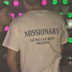 May include: A cream-coloured t-shirt with the word "MISSIONARY" printed in black, followed by "SO WE CAN KEEP ARGUING." The shirt is short-sleeved and appears to be made of cotton. The image is taken in a dimly lit environment.