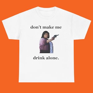 May include: White t-shirt with the text "don't make me drink alone." The shirt features an image of a person holding a gun. The person is wearing a pink shirt and a blue and white patterned scarf.