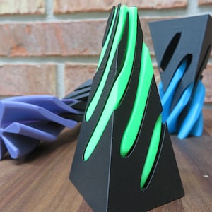 May include: A collection of abstract geometric sculptures in various colors. The central piece is a black pyramid with green curved accents. Other pieces include a blue, twisted form and a black and blue sculpture.
