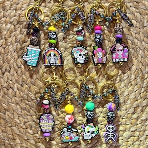 May include: A collection of keychains with gold-colored rings and decorative beads. Each keychain features a unique charm with phrases like "At Least My Trauma Made Me Funny" and "Horror Movies and Chill". The charms have colorful designs.