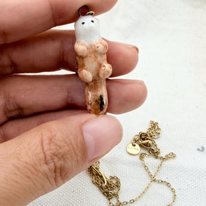 May include: A ceramic pendant shaped like a small otter with a white head and a light brown body, featuring a gold-toned metal chain. The otter has a small black nose and eyes, and the tail section includes a dark floral detail. A small round tag is attached to the chain.