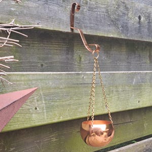 May include: A polished copper-coloured bowl suspended by three thin gold-toned metal chains attached to a matching copper-coloured wall-mounted hook. The bowl has a smooth, reflective surface and a rounded, hemispherical shape.