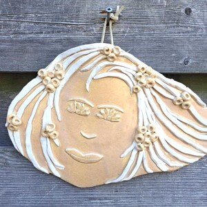 May include: A ceramic wall hanging depicting a stylized face with flowing hair and floral accents. The face is a light tan colour with white hair and floral details. The piece is suspended against a wooden backdrop.