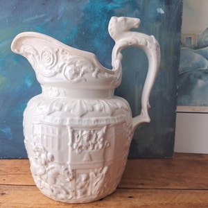 May include: White ceramic jug with detailed relief patterns. The jug has a spout, a curved handle with an animal head design, and relief patterns of flowers and architectural scenes. A decorative item.