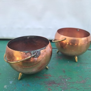 Peut inclure: Deux petits pots en cuivre avec des poignées et des pieds en laiton. Les pots sont ronds et ont une finition brillante. Ils sont posés sur une surface verte.