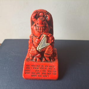 May include: A bright red figurine of a grinning devil character holding a small gold-coloured harp. The base features the text: YOU MAY NOT BE AN ANGEL AND I KNOW YOU'RE NOT A SAINT... BUT I LIKE YOU FOR WHAT YOU ARE AND NOT FOR WHAT YOU AIN'T.