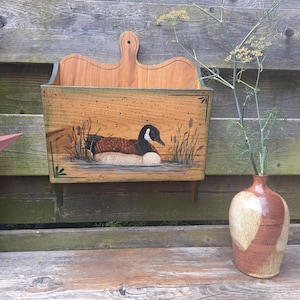 May include: A wall-mounted wooden magazine rack featuring a hand-painted scene of a Canada goose swimming in a pond with reeds. The rack has a natural wood grain finish, a curved top edge with a central hanging hole, and dark green painted accents.