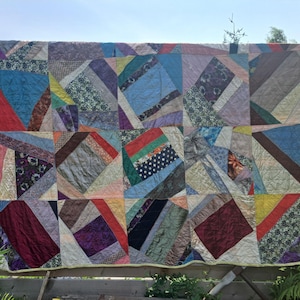 May include: A patchwork quilt with a variety of fabrics and patterns, including stripes, polka dots, and floral designs. The quilt is displayed outdoors, hanging on a wooden fence. The quilt's colours include blue, red, purple, and green.