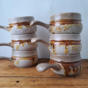 May include: A stack of ceramic soup bowls with handles. The bowls are beige with a brown and yellow glaze around the middle. The handles are curved and extend from the side of each bowl. The bowls are stacked on a wooden surface.