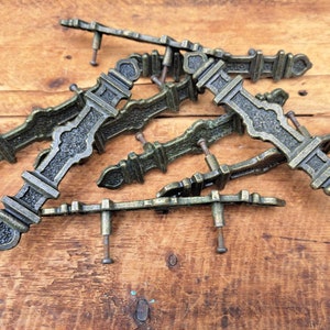 May include: Several antique bronze-effect drawer pulls. Each pull has an ornate, symmetrical design with decorative detailing. The pulls are arranged on a wooden surface, highlighting their aged appearance and intricate craftsmanship.