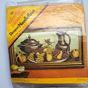 May include: A vintage needlepoint kit from Columbia Minerva, featuring a still life picture of a pitcher, bowl, grapes, and pears. The kit is labelled "Designer Needlepoint" and includes the text "Still Life Picture Approx. 24x12".