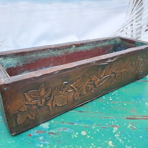 May include: A rectangular copper planter box featuring a raised relief pattern of ivy leaves along the front side. The metal surface displays a weathered brown finish with visible green oxidation and patina accents along the top edges and interior.