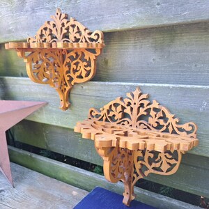 May include: Two intricately carved wooden wall shelves with a warm, natural finish. The shelves feature ornate, openwork designs and are mounted on a weathered wooden fence. The shelves are approximately 30 cm wide.