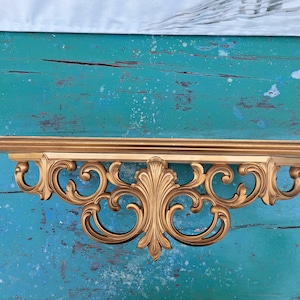 May include: A gold-colored, ornate shelf bracket with a decorative, scroll-like design. The shelf bracket is mounted on a teal-colored wall.