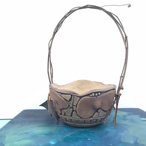 May include: A brown ceramic planter with a woven vine handle. The planter has a textured surface and is decorated with a pattern of circles and squares.