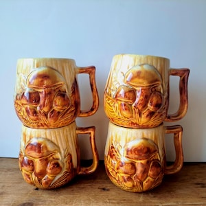 May include: Four ceramic mugs with a mushroom design. The mugs are stacked in pairs, showcasing a detailed relief of mushrooms in shades of brown and tan. Each mug has a large handle and a textured, wood-like appearance.