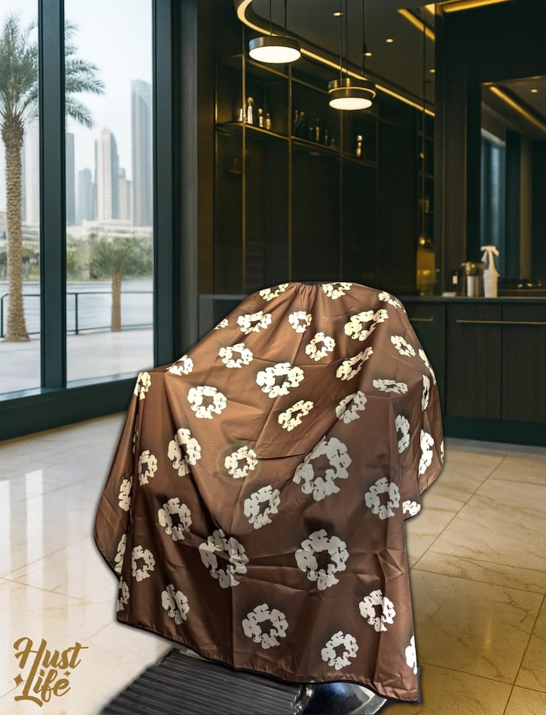 K&ouml;nnte beinhalten: Ein brauner Friseurumhang mit wei&szlig;em Blumenmuster bedeckt einen Stuhl. Der Umhang besteht aus einem glatten Stoff und soll die Kleidung beim Haareschneiden sch&uuml;tzen. Das Hust Life-Logo ist unten links sichtbar.