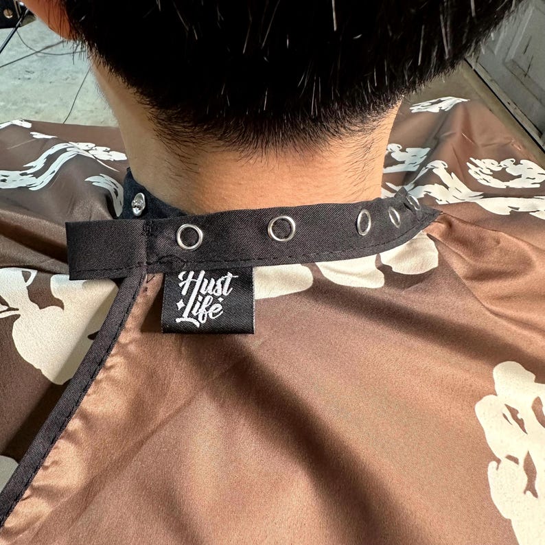 K&ouml;nnte beinhalten: Braune und wei&szlig;e Friseurumhang mit floralem Muster, schwarzem Kragen und dem Text "Hustle Life" auf einem schwarzen Etikett. Der Umhang hat Metall&ouml;sen und einen schwarzen Riemen zum Verschlie&szlig;en.